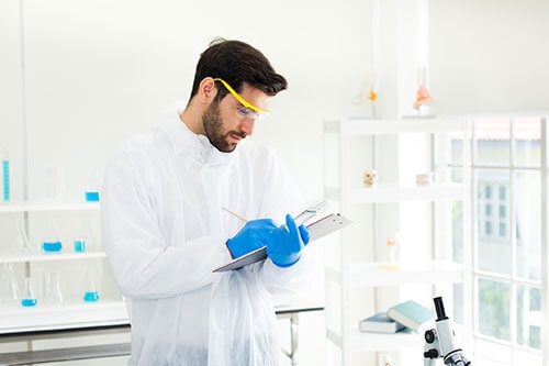 A man in lab coat and goggles writing on paper.