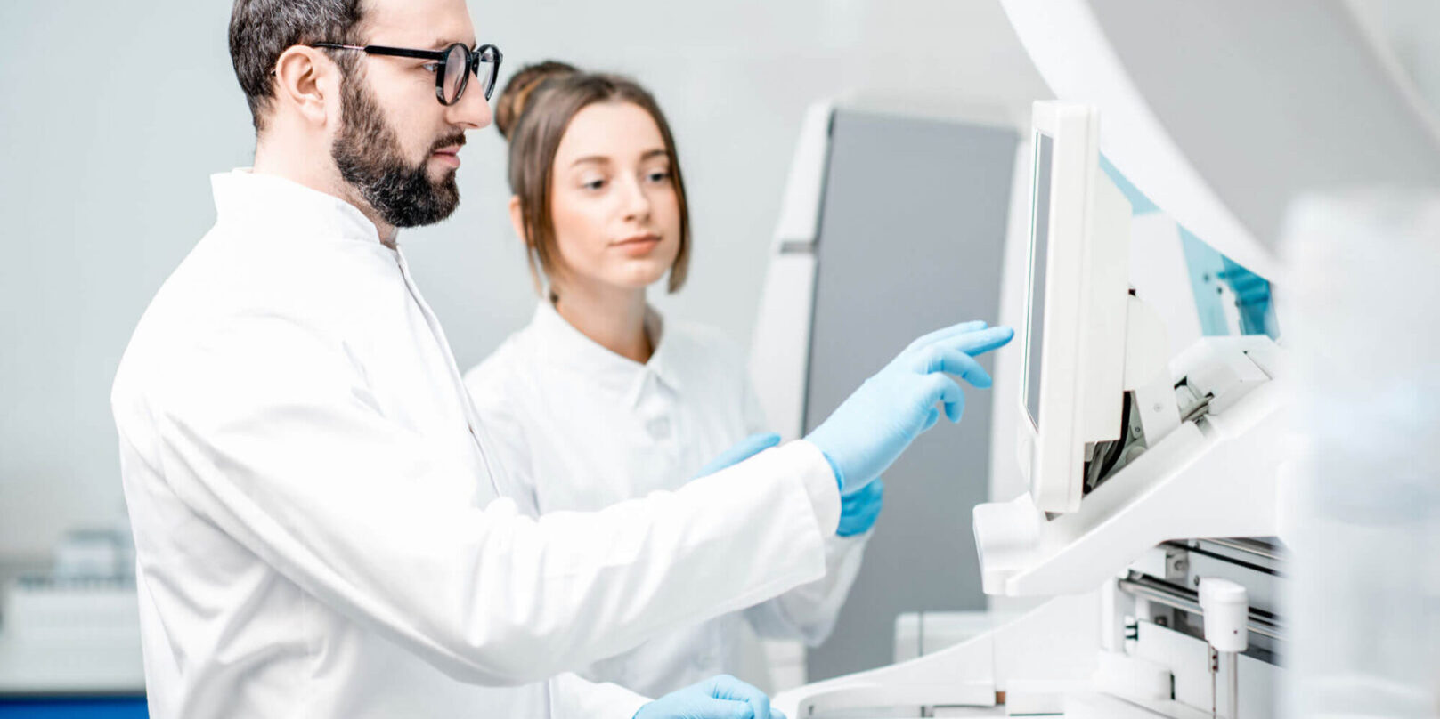 GettyImages-946219890-copy Two people in lab coats and gloves looking at a computer.